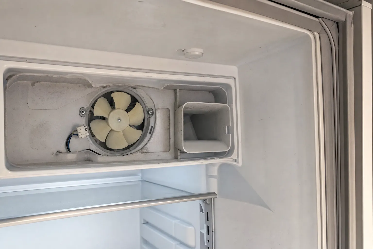 Inside an empty white compartment with the upper duct cover off to show a fan, the door gasket running down the frame edge